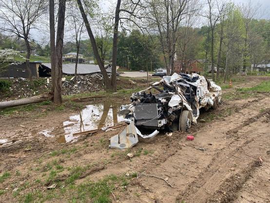 White vehicle damaged beyond recognition in foreground. Collapsed building in the distance