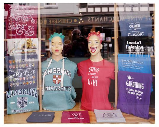The image displays a store window featuring a variety of merchandise themed around Cambridge University. There are two mannequin heads with brightly colored hair (one blue and one red) wearing t-shirts with humorous statements. On the left, apparel that showcases the Cambridge University logo, such as shirts and hoodies, is visible. On the right, there are t-shirts and colored totes with quirky sayings like "Gym?? I thought you said Gin!" and other humorous designs. The setup appears to be aimed at tourists or students, offering both university-branded merchandise and novelty items.