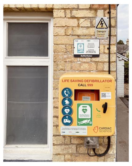 This image shows a publicly accessible defibrillator unit mounted on a brick wall next to a window. The defibrillator is housed in a bright yellow case labeled "LIFE SAVING DEFIBRILLATOR CALL 999" with instructions and icons indicating how to perform CPR and use the AED (Automated External Defibrillator). There are also several safety signs, including a warning about overhead wires and a notice explaining that the defibrillator is monitored by CCTV and that its misuse constitutes a severe offense. The brand "Cardiac Science" is visible at the bottom of the defibrillator unit.