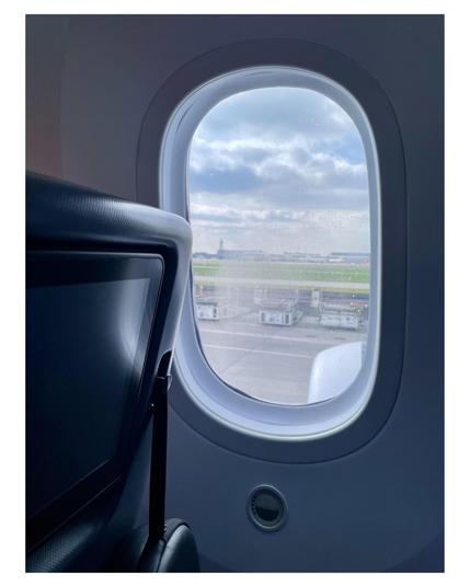 The image shows the view from the window seat of an airplane. The window frame is visible, and through the glass we can see the tarmac of an airport. There are various ground service equipment pieces, like trolleys and vehicles, visible outside. The sky above is partly cloudy and bright, suggesting it could be a sunny day. The general ambiance is calm and typical of what one might see while sitting on a parked or taxiing airplane waiting for takeoff or just after landing.