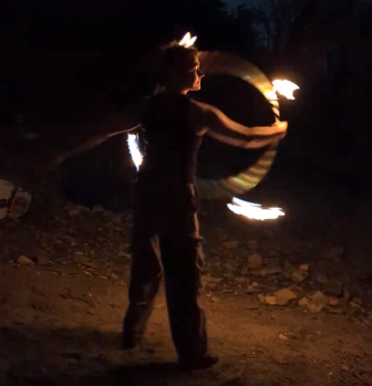 A man spinning a fire hoop on his arm, standing on a rocky terrain at night. The silhouette of his face  is illuminated by the glow of the four flames around the hoop.