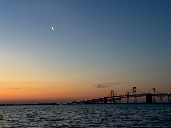 The bay bridge at sunrise with a crescent moon setting. Busy morning traffic lights are visible on the bridge.