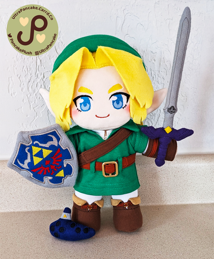 A 12 inch tall plush of Link from Ocarina of Time. He has short blonde hair that's parted down the middle. He's wearing a green tunic, green boots and a green hat. He's holding the Master Sword in his left hand and the Hylian Shield in his right. At his feet sits a tiny plush Ocarina.