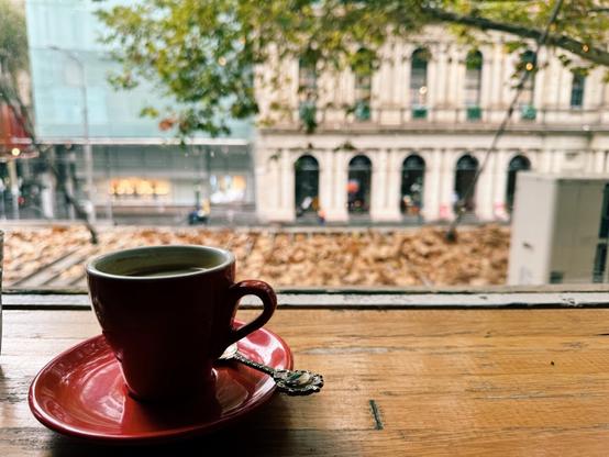 A red cup of coffee sits on a red saucer with a spoon, on a ledge just inside of a window. Outside, autumn leaves are scattered across an awning just below the window; beyond is a tree, and some buildings. It’s a bit cold and wet.