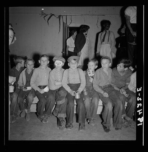 The image is a black and white photograph depicting nine boys sitting side by side on what appears to be a bench or platform. They are dressed in various styles of clothing, some wearing overalls with suspenders and others shirts and pants. Most of the boys have cups held near their mouths, suggesting they might be waiting for refreshments at an event like Halloween. Several of them wear costumes; one has a mask resembling a cartoonish character on his face, while another sports a hat with white squares or checks patterned across it.

Behind the children stands several adults who seem to be overseeing the group, possibly staff members from the Shafter Migrant Camp in California where this event was likely taking place. The atmosphere seems casual and communal as they wait for further instructions on what's about to happen during their gathering.