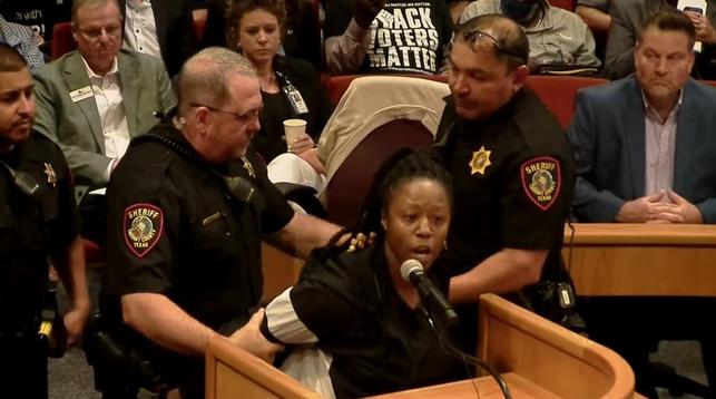Janell Johnson [a black woman speaking at a podium/lectern], the sister of a man killed by guards inside the Tarrant County jail, was forcibly removed by deputies [both white and POC] during a county meeting on June 4, 2024. (Screenshot/Tarrant County) 

In the background are several rows of public seating for the meeting, with one shirt reading "Black Voters Matter."