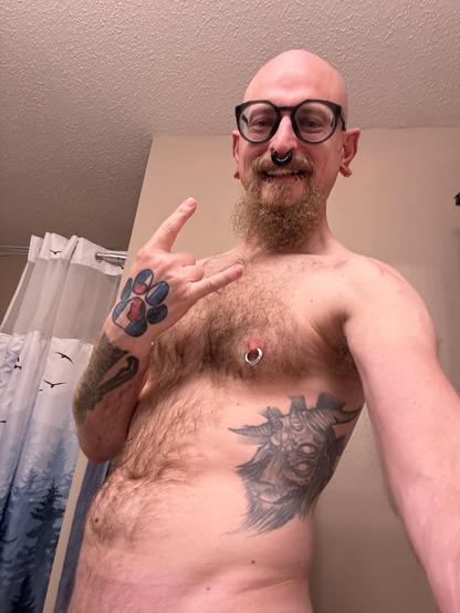 A shirtless person with a beard and glasses is posing in a bathroom. They have multiple tattoos, facial piercings, and are throwing up the horns with their right hand. The setting features a shower curtain in the background.
