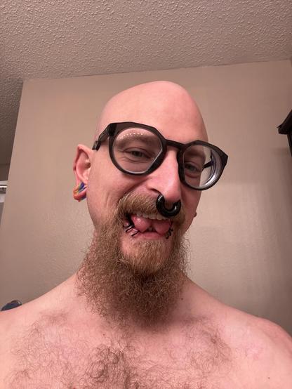 A bald person with a full goatee smiles sticking their forked tongue out at the camera, wearing large, distinctive glasses. They have multiple facial piercings, including a large gauge septum ring and 6 rings in their lip, and they are shirtless in a bathroom setting.