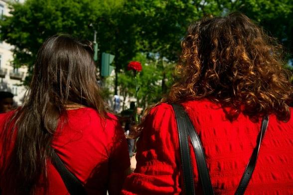 Two people with long hair and wearing red tops stand outdoors, one holding a red flower.