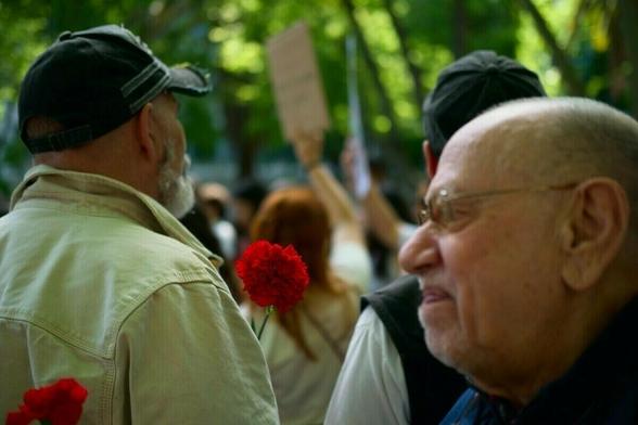A group of people is gathered outdoors, with one person holding a red carnation and others visible in the background, including a blurred protest sign.
