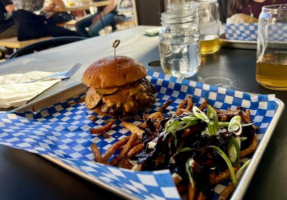 A tray with a cheeseburger and fries topped with sauce and green onions sits on a table alongside drinks in jar glasses.