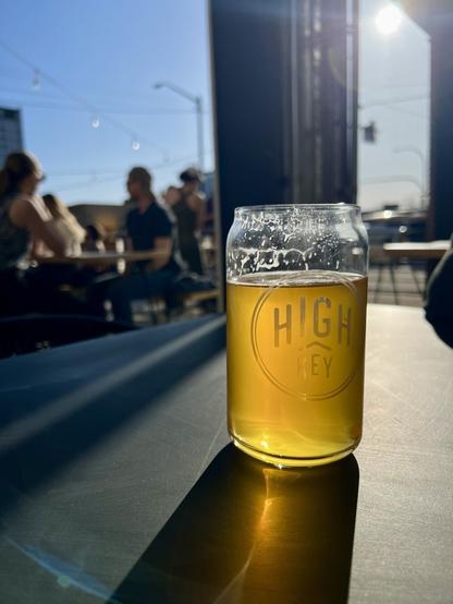 A glass of beer with the logo "High Key" sits on a table outdoors, with people and a sunny sky in the background.