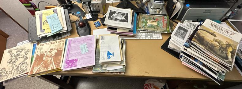 A panoramic shot of a desktop, currently looking very cluttered due to the eight stacks of SFF convention program books, flyers, and other assorted ephemera piled on top of and around the barely visible computer keyboards, tablet, and trackball that normally occupy this space.