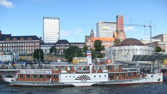 Seitenrad-Dampfer mit weißem Rumpf, weißen Radkästen und Naturholz-Aufbauten. Im Hintergrund die St. Pauli-Landungsbrücken und verschiedene hohe Gebäude.