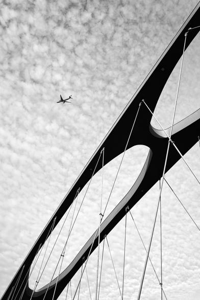 A filigree bridge construction taken from the frog's perspective. The background is the cloudy sky in which a passenger airplane is flying.