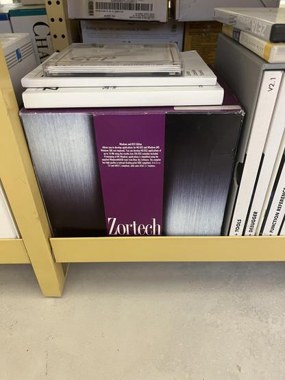 A shelf displays a boxed software product labeled "Zortech," which appears to be related to development for MS-DOS and Windows. Several other software boxes are stacked next to it. The box features a purple color scheme with a metallic background.