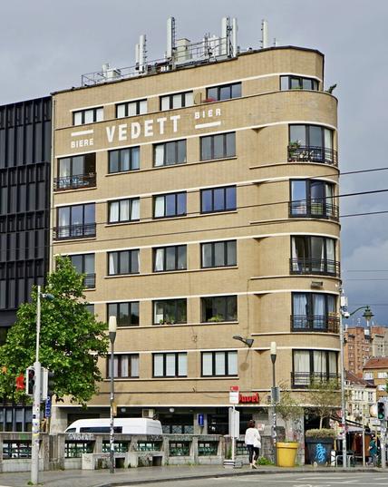 Grand immeuble moderniste en brique jaune faisant l’angle d’un quai. Il compte six étages et porte un marquage commercial des bières Vedett en lettres blanches. L’angle de l’immeuble est arrondi, évoquant le style paquebot des années 30.