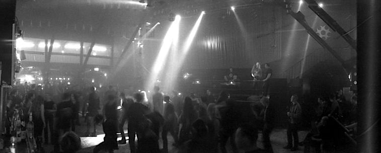 A panorama shot on analogue black-and-white film in a discotheque. It shows the dance floor on which people can be seen dancing in the shadows. The room is supported by wooden struts - a few beams of spotlights illuminate the scene