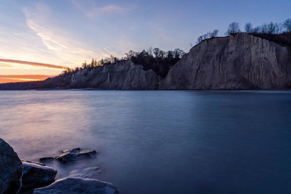 Twilight view of the towering Scarborough Bluffs in Toronto, with smooth lake water in the foreground and a soft orange glow on the horizon.