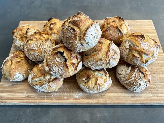 Auf einem dunklen Holztisch liegt ein Holzbrett. Darauf liegen zahlreiche frisch gebackene Brötchen mit rustikalem Ausbund.