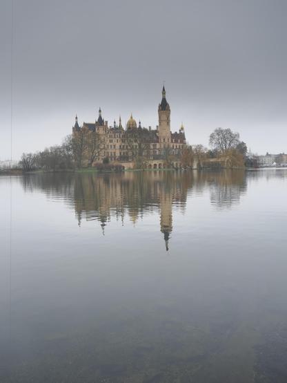 The image depicts a large, historic castle with numerous spires and towers, situated on a piece of land surrounded by a body of water. The castle is reflected in the calm water, and there are trees and muted vegetation around it, under a cloudy sky.