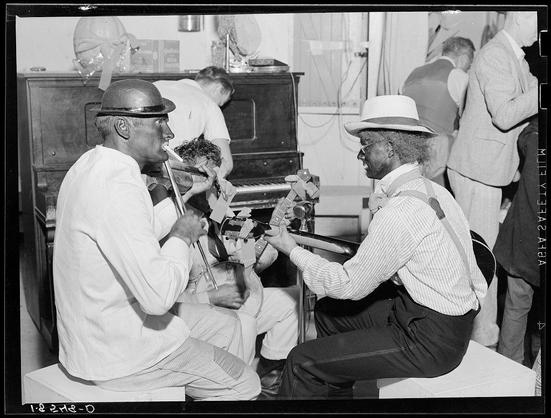 The image is a black and white photograph depicting two musicians playing brass instruments in front of an upright piano. They appear to be performing or practicing together, with one musician on the left holding a trumpet-like instrument close to his mouth as if he's about to play it, while the other man on the right holds what appears to be a trombone.

Both individuals are seated and dressed casually; they wear hats and shirts but no jackets. The person playing the horn has a hat with some sort of emblem or design in front, whereas the individual holding the trombone wears glasses and suspenders over his striped shirt and dark trousers.

The setting seems informal, possibly part of an impromptu performance during a social event such as a party or gathering. In the background, other individuals can be seen engaging with their own activities: one person is bent down near another seated piano player who's focused on playing it; others are standing around conversing and enjoying themselves.

The image provides insight into community life and entertainment from an earlier time period in California, specifically Shafter camp for migrants during Halloween.