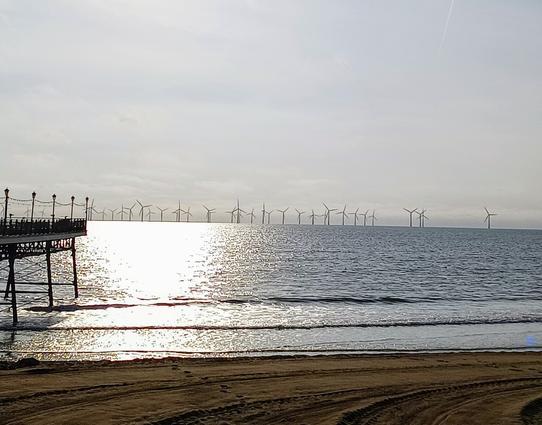 A shot wind turbines taken from a beach. The wind turbines are out at sea in the distance, there is a pier on the left hand side of the photo.

The sun is low in the sky, reflecting off the ocean.