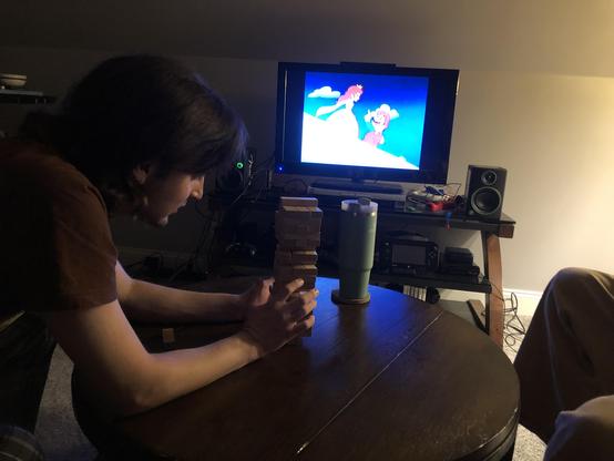 A young man carefully removing a block from a Jenga tower. The Super Mario Bros. Super Show is on in the background.