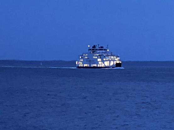 a ship passing in the night. a car ferry with all the deck spaces brightly illuminated so it stands out against the deep blue sea and sky