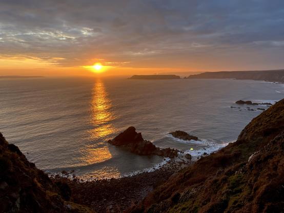 Sunset over a calm sea. In the foreground rocks beside the shore. In the horizon a headland and an island.