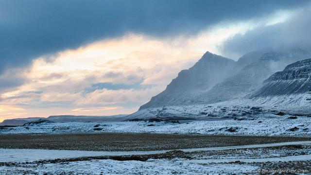 A colour photo of a coastline covered in ice and snow. The waterside is craggy and spikes of dark rock show through. On the right is a tall pyramidal mountain, silhouetted and draped in scarves of cloud. The sky is heavily overcast and grey, but a window is opened in it through which the orange of dawn is visible.