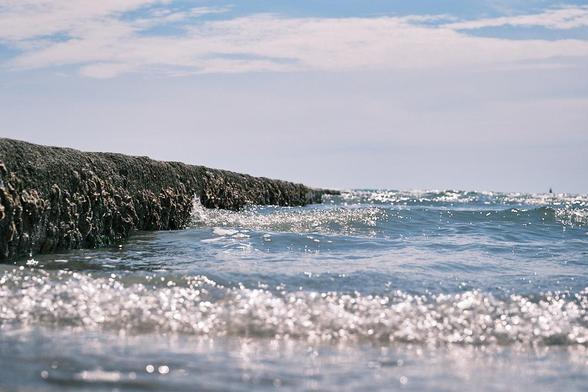 Sunlight sparkles on blue sea waves hitting a rough, dark breakwater. Shot from a low perspective.