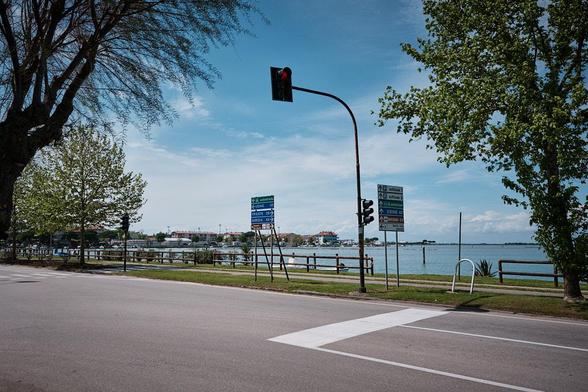 Road intersection in Grado, Italy, with a red traffic light and signs for Trieste, Udine, and Gorizia. Water and town visible behind under a blue sky.