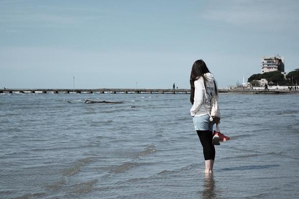 My girlfriend stands ankle-deep in the sea, facing away towards a distant pier, holding a bag with her shoes hanging from it.