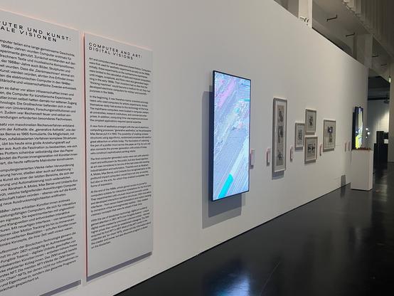 Photo of a gallery space with printed and screen-based abstract artworks and info panels mounted on a long white wall. One of the pieces (titled "De/Frag", by the author) is shown on a large 75" screen mounted in portrait format, its light reflecting on the shiny black floor. Image credit: Clara Runge
