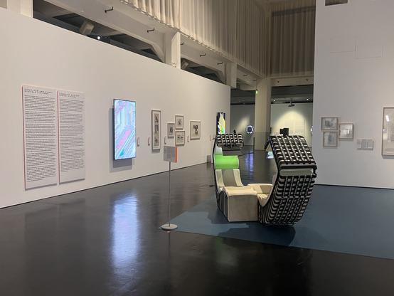Photo of a large gallery space with artworks and info panels mounted on a long white wall and a sculpture in the center of the space. One of the pieces (titled "De/Frag", by the author) is shown on a large 75" screen mounted in portrait format, its light reflecting on the shiny black floor. Image credit: Clara Runge