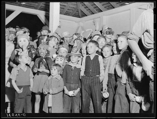 The black and white photograph captures a group of children gathered together at what appears to be an event or party. Many of the kids are wearing various costumes, suggesting that this could be around Halloween time. The attire ranges from simple overalls and dresses to elaborate outfits with accessories such as hats adorned with faces or props resembling different objects.

The setting looks like an outdoor area possibly within a larger structure or covered pavilion due to visible beams overhead. There's also another individual partially in the frame, seemingly assisting the children. One child is holding what seems like a cup from which they might be drinking, and others have their hands on hips or pockets as if waiting for something.

The image carries with it an aura of community spirit among these young attendees who seem engrossed in the event's activities despite not being able to fully see each other. The overall composition conveys a sense of nostalgia and communal joy experienced during childhood gatherings, particularly around holidays like Halloween which are often celebrated through costume parties at migrant camps or rural communities where such events might have been significant social occurrences.

Additional context is provided by the link given in your question: "Halloween party at Shafter migrant camp, California." This indicates that this photograph was likely taken in a migrant worker's community d [...]