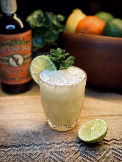 light yellow cocktail with fresh lime wheel and mint sprig garnish in a faceted glass sitting on an etched wooden cutting board next to a half lime with citrus and a bottle of Doctor Bird pot still overproof rum in the background