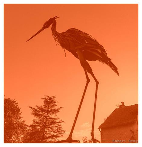 A colorised image of a metal sculpture of a heron silhouetted against the sky with trees  and the roof of a farmhouse in the background.