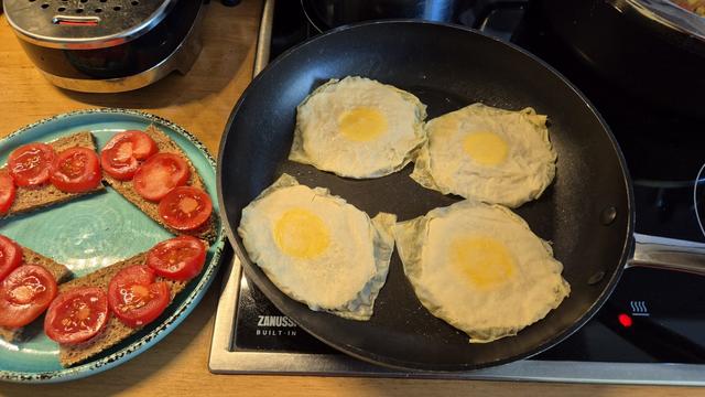 In einer Pfanne werden gerade vier vegane Spiegeleier gebraten, links neben der Pfanne steht bereits ein vorbereiteter Teller mit Brot, das mit Tomatenscheiben belegt ist.
