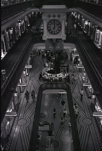 A grainy B&W wide shot of the Queen Victoria Building in Sydney, Australia. The shot is from the top of the building, looking down at about 45 degrees. People walk about the various stores, storefronts and signage just visible in the sides. A clock looms over it all.