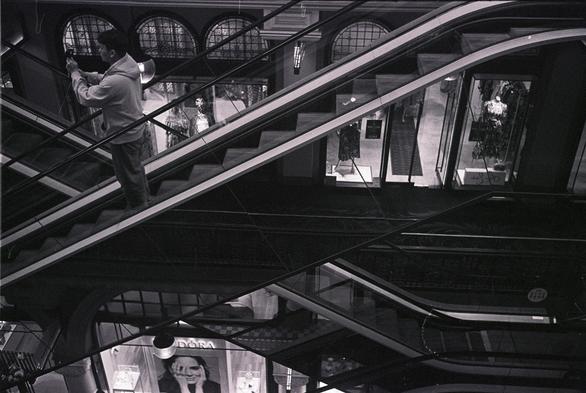 A grainy B&W photo of two single-width escalators in the Queen Victoria Building crossing each other. They are reflective on the sides, showing more of the building. A man is on one of the escalators, holding a DJI camera or similar as he rides the escalator.