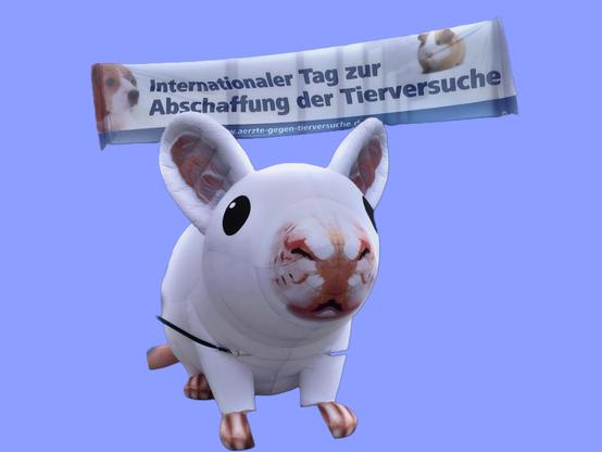 Eine aufblasbare Maus sitzt vor einem blauen Hintergrund und darüber ein Banner mit "Internationaler Tag zur Abschaffung der Tierversuche"