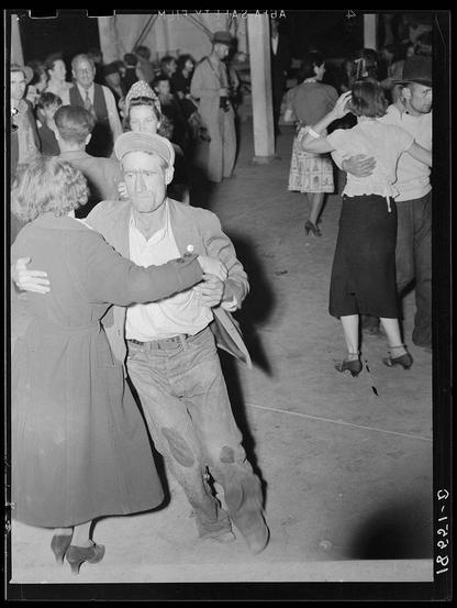 The provided photograph is a black and white image depicting what appears to be an event or gathering, possibly from the early to mid-20th century. The scene shows several individuals engaged in dancing on a spacious floor that suggests it might have been held indoors. People are dressed in casual attire; women wear dresses with various styles while men don shirts paired with trousers and some sporting hats. One man at the forefront is notably older, wearing a cap, overcoat, and showing signs of wear or distress such as patched pants.

The environment has an atmosphere that could be interpreted as communal but also carries traces of hardship, hinted by the clothing's condition. The photo seems to capture candid moments with no apparent posing, emphasizing a real-life scenario rather than staged event photography. There is no clear indication of age group differences within this single image; however, it depicts adults engaging in social activities typically associated with leisure or celebration.

The watermark and date on the photograph indicate its historical context: "Dorothea Lange - 1942-03". The specific mention of 'Aid A' alongside a location tag could point to some organizational involvement. Overall, this image is rich in detail that suggests it was taken during an informal social event possibly among migrant workers or attendees at a local community gathering given the historical context and attire style.

The  [...]