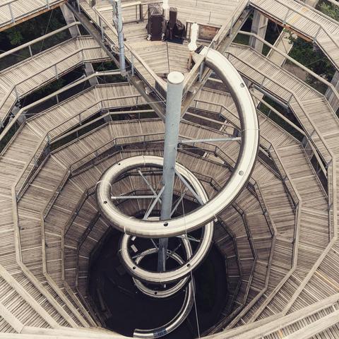 Das Bild zeigt eine beeindruckende Struktur, die aus einem zylindrischen Turm mit einem spiralförmigen Aufgang besteht. Im Zentrum des Turms befindet sich eine große, spiralförmige Metallrutsche, die sich mehrmals im Zentrum des Turms hinunter windet. Die Rutsche ist silbern und glänzt im Licht. Am oberen Ende des Turms befinden sich einige Geräte und eine Plattform, von der aus man die gesamte Struktur überblicken kann. Der Turm ist Teil eines Baumwipfelpfades. Die Struktur ist sowohl funktional als auch ästhetisch ansprechend, mit einer Mischung aus natürlichen und modernen Materialien.