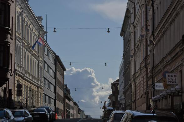 A street with cars parked on either side, tall (not modern) buildings on either side and in the horizon, a blue sky with silver-lined clouds