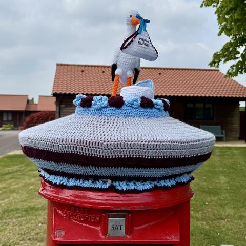 Postbox with a knitted topper showing a stork carrying a baby in a blanket and a crib underneath it.