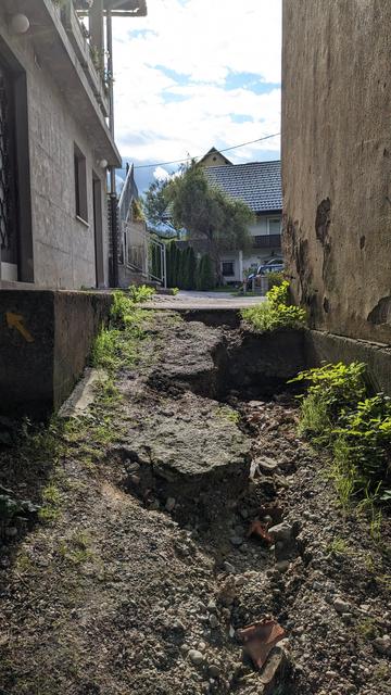Hochkant in Farbe aus der Froschperspektive. Blick auf einen trockenen, erdigen Streifen (ca 1,20 m breit) zwischen zwei Häusern mit Blick auf eine Straße. Von den Häusern sieht man nur die Seitenwände. Der Erdstreifen fällt zum Betrachter hin ab. Wasser hat sich einen tiefen Lauf in die Erde gegraben, Steine und Schutt wurden freigespült. An den Rändern des Erdstreifens wachsen niedrige Wildkrautpflanzen. Es ist sonnig mit Wolken.