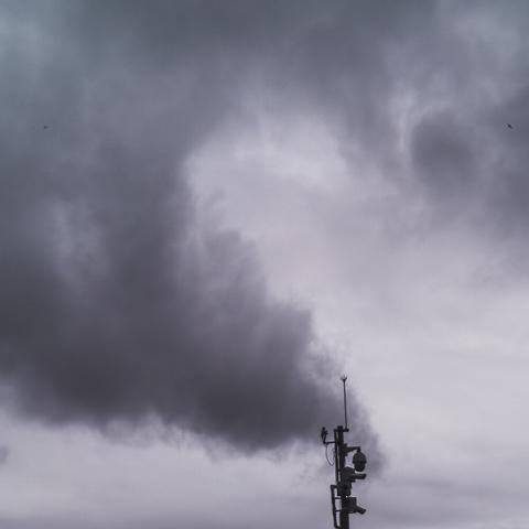 A photo of a bunch of street cameras on a pole in the bottom right of the image, from which it appears as if smoke is billowing out in a spiral above it. In reality, it is just  dark clouds in the background which happen to align with the cameras.