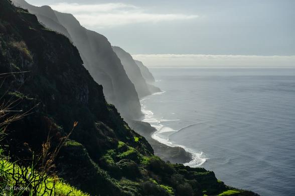 A misty coastal landscape featuring steep green cliffs along the shoreline, with the ocean reflecting soft light and gentle waves.
